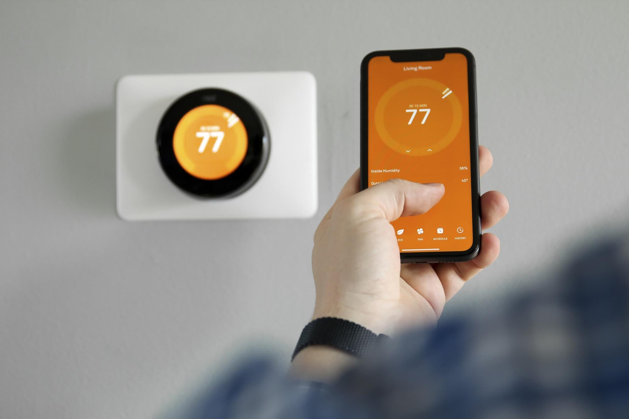Person holding smart phone with display mirroring the smart thermostat temperature behind it on the wall